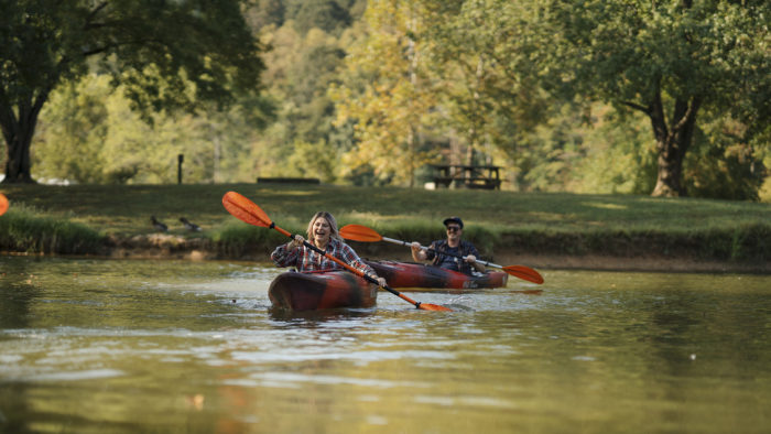 Smiling couple kayaking on a calm river near Huntington, West Virginia, surrounded by trees and parkland — a popular outdoor activity featured by the Huntington Area CVB for adventure seekers and nature lovers
