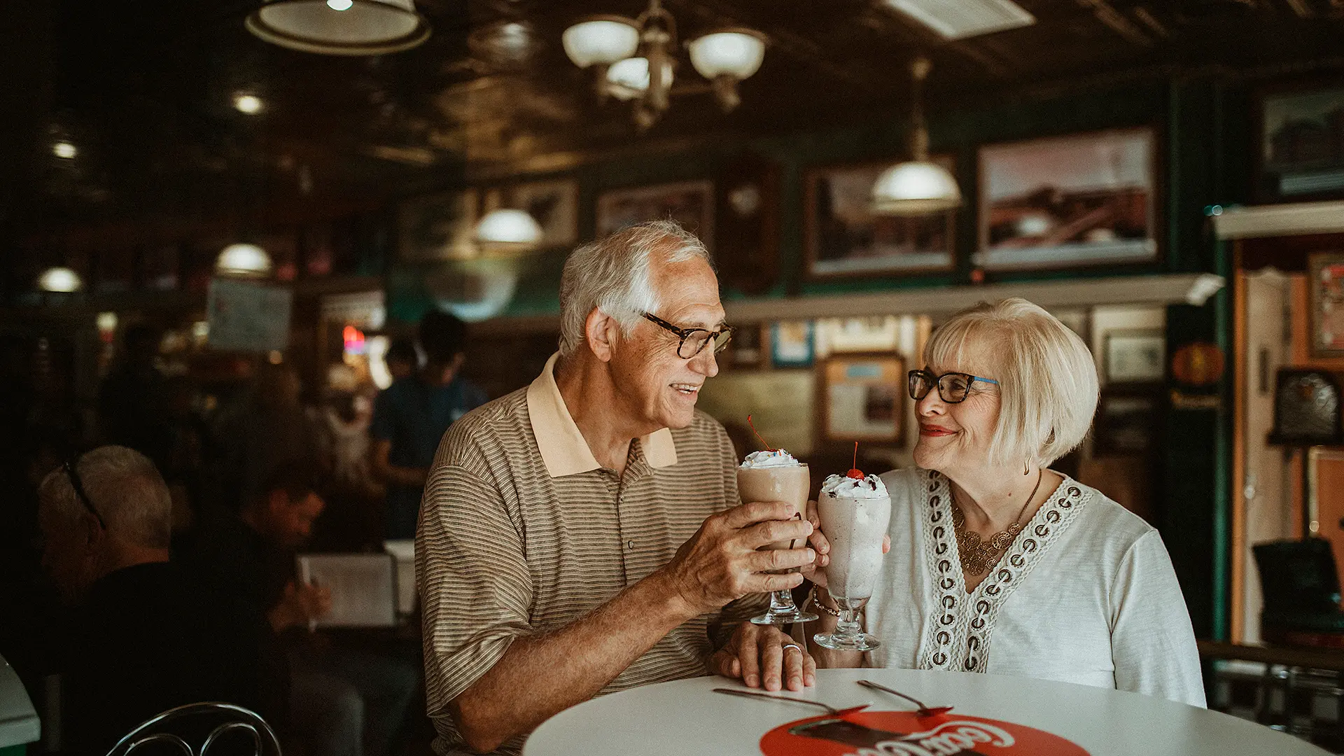 Smiling couple sharing milkshakes at a retro-style diner, surrounded by vintage decor and a lively atmosphere — a cozy and nostalgic setting perfect for classic desserts and memorable moments.