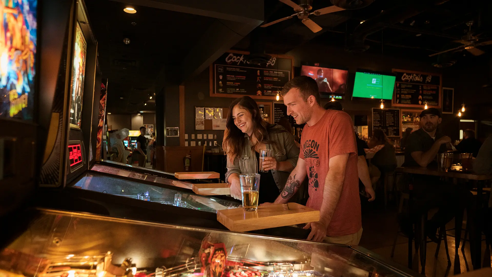 Two adults playing pinball and holding drinks inside a lively bar with arcade games